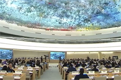 [ai] A large assembly hall with rows of delegates seated at wooden desks, some facing a screen displaying images. The ceiling features a colorful mural. The atmosphere appears formal, likely during an international conference or meeting.